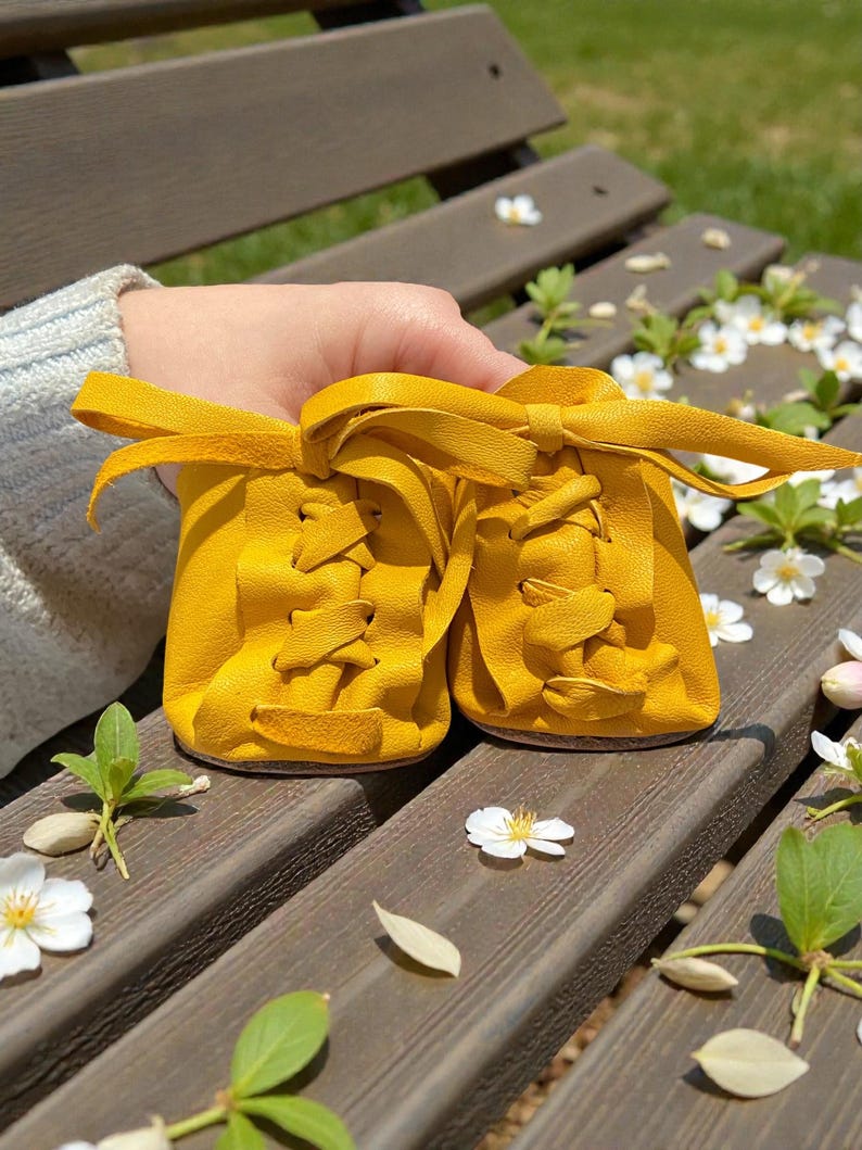 May include: A pair of bright yellow baby shoes with laces and a bow. The shoes are made of soft leather and are sitting on a wooden bench. The background includes white flowers and greenery.