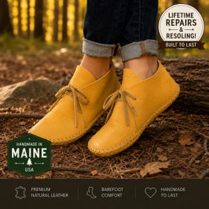 May include: A pair of mustard yellow leather moccasins with thin laces and visible whipstitch detailing along the sole. The shoes are worn with cuffed dark blue jeans. Text overlays include "HANDMADE IN MAINE" and "LIFETIME REPAIRS & RESOLING!"
