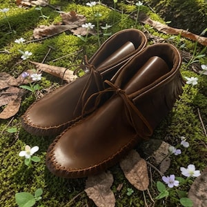 Puede incluir: Un par de mocasines de cuero marr&oacute;n con un detalle de costura de l&aacute;tigo a lo largo de la punta redondeada, un borde de flecos alrededor del cuello del tobillo y cordones de cuero finos atados en un lazo en la parte delantera.