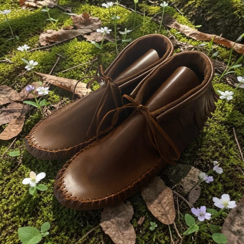 Puede incluir: Un par de mocasines de cuero marr&oacute;n con un detalle de costura de l&aacute;tigo a lo largo de la punta redondeada, un borde de flecos alrededor del cuello del tobillo y cordones de cuero finos atados en un lazo en la parte delantera.