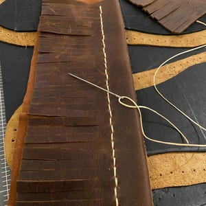 May include: Close-up of leather craft in progress. A piece of brown leather with fringed edges is being hand-stitched with cream-colored thread. A needle is inserted through the leather, with other leather pieces in the background.