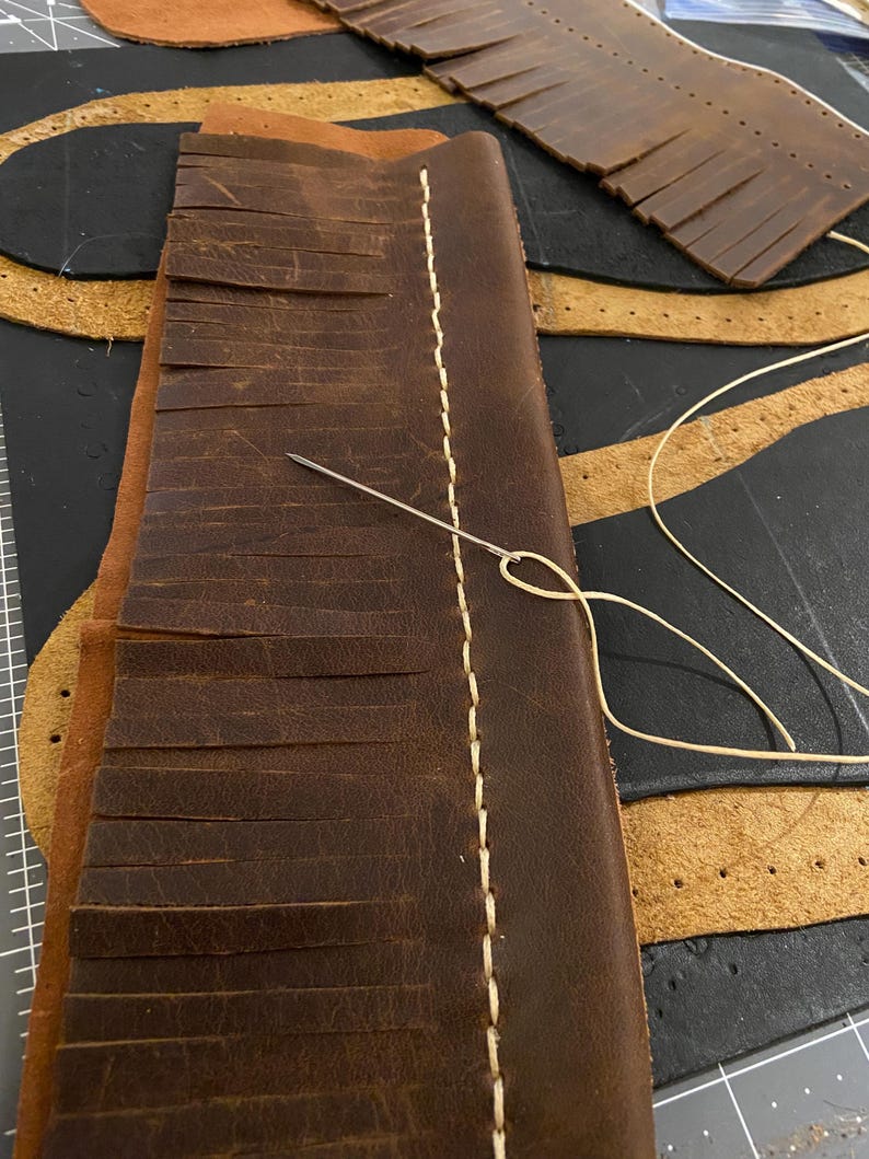 May include: Close-up of leather craft in progress. A piece of brown leather with fringed edges is being hand-stitched with cream-colored thread. A needle is inserted through the leather, with other leather pieces in the background.