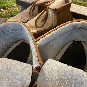 May include: Close-up of a pair of moccasin boots with a light brown exterior and a light blue interior. The boots have a fringe detail and leather laces. The background is blurred, showing a wooden bench and greenery.