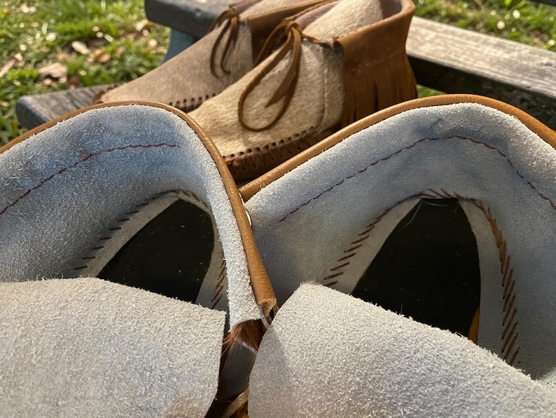 May include: Close-up of a pair of moccasin boots with a light brown exterior and a light blue interior. The boots have a fringe detail and leather laces. The background is blurred, showing a wooden bench and greenery.