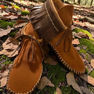 May include: A pair of brown suede moccasin boots with a fringed collar and leather laces. The boots have white stitching around the soles and are set against a backdrop of fallen leaves and moss.