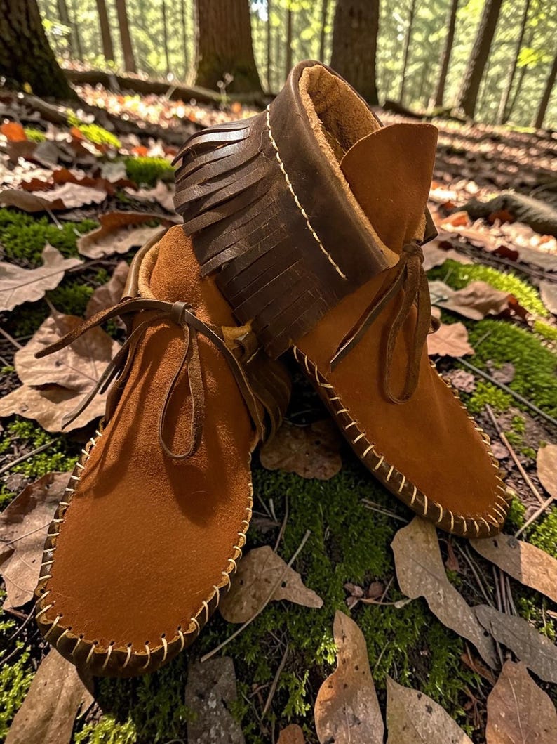 May include: A pair of brown suede moccasin boots with a fringed collar and leather laces. The boots have white stitching around the soles and are set against a backdrop of fallen leaves and moss.