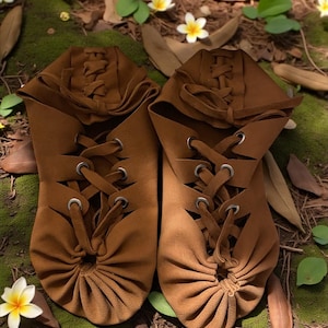 May include: A pair of brown leather moccasin-style shoes with intricate lacing and eyelets. The shoes have a gathered design at the toe and are set on a bed of moss, leaves, and small white and yellow flowers.