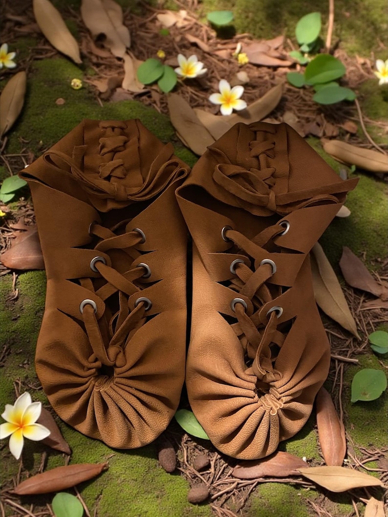 May include: A pair of brown leather moccasin-style shoes with intricate lacing and eyelets. The shoes have a gathered design at the toe and are set on a bed of moss, leaves, and small white and yellow flowers.