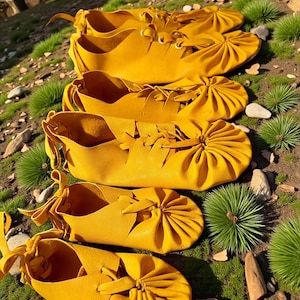 May include: A collection of handmade, yellow leather moccasins. The shoes are arranged in a row, showcasing their unique design with laces and gathered details. The moccasins are displayed on a bed of green grass and small rocks.