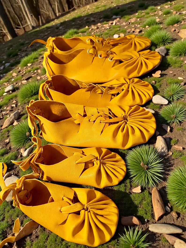 May include: A collection of handmade, yellow leather moccasins. The shoes are arranged in a row, showcasing their unique design with laces and gathered details. The moccasins are displayed on a bed of green grass and small rocks.