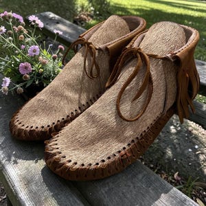 Puede incluir: Un par de mocasines de color tostado con una parte superior de piel texturizada, ribetes de cuero marr&oacute;n, detalles de costura en la puntera y cordones de cuero finos atados en la parte delantera. Los zapatos incluyen flecos decorativos en el tal&oacute;n.