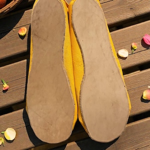 May include: A pair of yellow ballet flats with light brown soles. The shoes are made of a textured material and are displayed on a wooden surface. The image is taken from a low angle, showcasing the soles of the shoes.