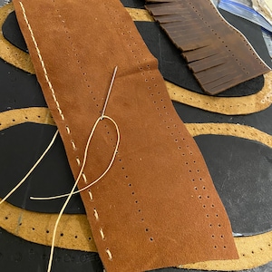 May include: A close-up of leather crafting materials. A brown leather piece with pre-punched holes is being hand-stitched with a needle and cream-colored thread. Other leather pieces in various shapes and colors are in the background, suggesting a leatherwork project.