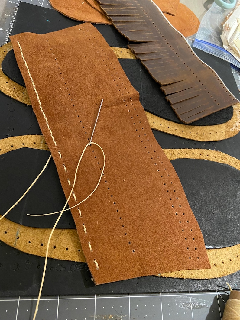 May include: A close-up of leather crafting materials. A brown leather piece with pre-punched holes is being hand-stitched with a needle and cream-colored thread. Other leather pieces in various shapes and colors are in the background, suggesting a leatherwork project.