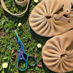 May include: Tan leather moccasin-style shoes with gathered detailing and a lace-up design. A pair of iridescent scissors and a tan leather strap are also visible, all set against a backdrop of green foliage and small yellow flowers.