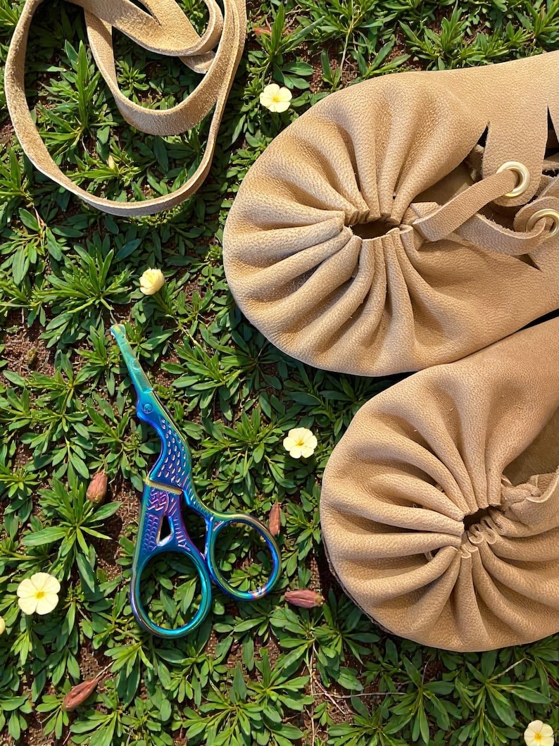 May include: Tan leather moccasin-style shoes with gathered detailing and a lace-up design. A pair of iridescent scissors and a tan leather strap are also visible, all set against a backdrop of green foliage and small yellow flowers.