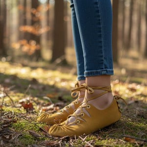 May include: Mustard yellow moccasin-style shoes with ankle ties and laces. The shoes are worn with blue jeans. The image is taken outdoors in a forest setting with green moss and fallen leaves on the ground.