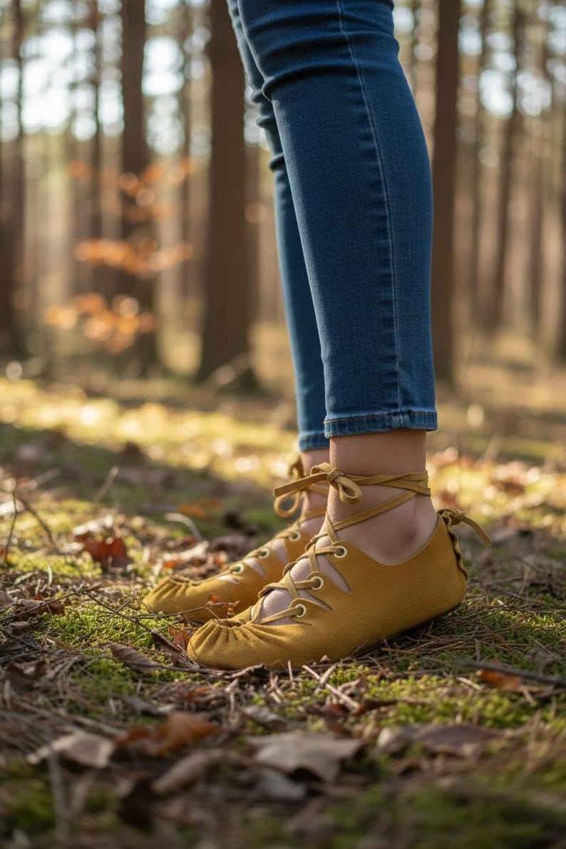 May include: Mustard yellow moccasin-style shoes with ankle ties and laces. The shoes are worn with blue jeans. The image is taken outdoors in a forest setting with green moss and fallen leaves on the ground.