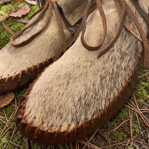 May include: Close-up of a pair of handmade moccasin-style shoes. The shoes feature a light brown and white speckled fur upper, dark brown leather soles, and are stitched with orange thread. Brown leather laces are tied at the top.