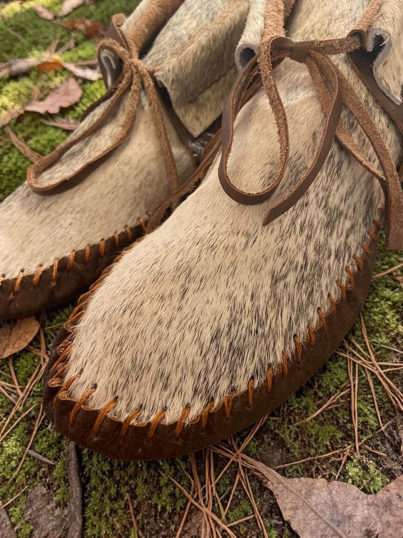May include: Close-up of a pair of handmade moccasin-style shoes. The shoes feature a light brown and white speckled fur upper, dark brown leather soles, and are stitched with orange thread. Brown leather laces are tied at the top.