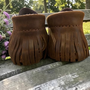 Puede incluir: Un par de mocasines de cuero marr&oacute;n con un detalle de flecos alrededor del tal&oacute;n. El calzado est&aacute; construido con costuras visibles a lo largo del borde superior de los flecos, mostrando un acabado mate y texturizado en el cuero.