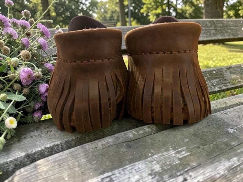 Puede incluir: Un par de mocasines de cuero marr&oacute;n con un detalle de flecos alrededor del tal&oacute;n. El calzado est&aacute; construido con costuras visibles a lo largo del borde superior de los flecos, mostrando un acabado mate y texturizado en el cuero.