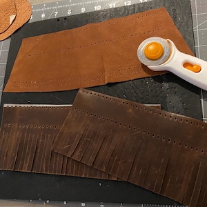 May include: A leather crafting scene with brown leather pieces, a rotary cutter, pins, and scissors. The leather pieces are perforated and some have fringe. The rotary cutter is white and orange. A purple container holds the pins.