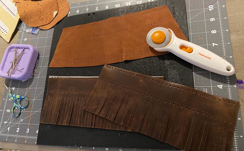 May include: A leather crafting scene with brown leather pieces, a rotary cutter, pins, and scissors. The leather pieces are perforated and some have fringe. The rotary cutter is white and orange. A purple container holds the pins.