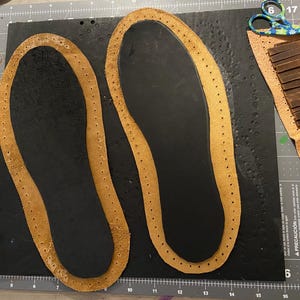 May include: Two brown leather insoles with stitched edges sit on a black surface. Additional leather pieces and a pair of scissors are visible, suggesting a craft project. The insoles are for shoes or sandals.