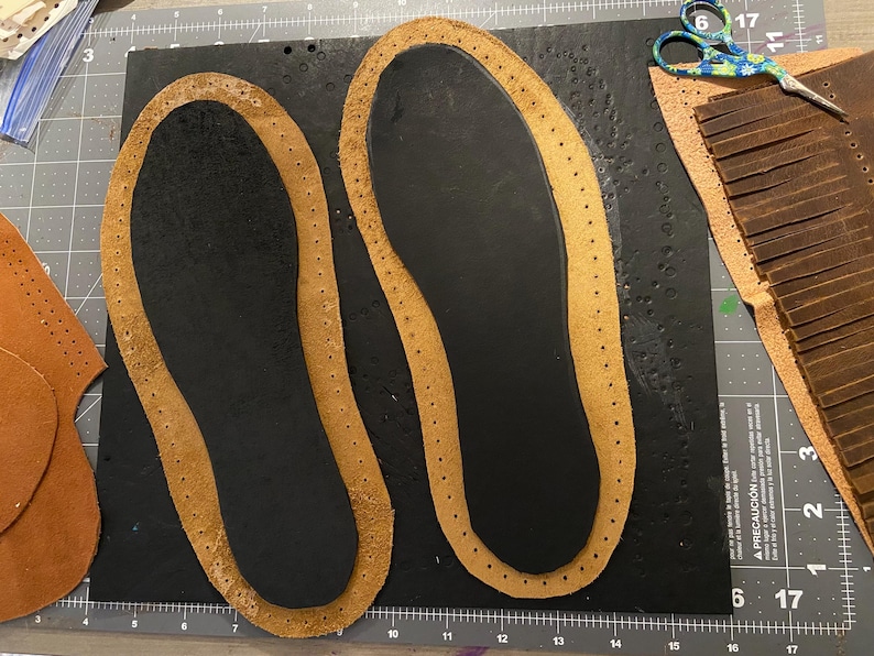 May include: Two brown leather insoles with stitched edges sit on a black surface. Additional leather pieces and a pair of scissors are visible, suggesting a craft project. The insoles are for shoes or sandals.