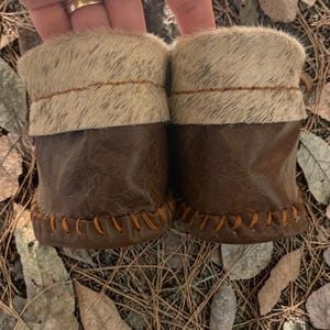 May include: A pair of baby moccasins with brown leather bottoms and a light-colored fur cuff. The moccasins are hand-stitched with orange thread. The background is a bed of leaves and pine needles.