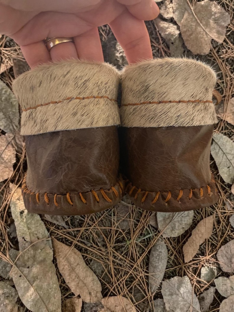 May include: A pair of baby moccasins with brown leather bottoms and a light-colored fur cuff. The moccasins are hand-stitched with orange thread. The background is a bed of leaves and pine needles.
