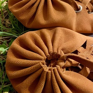 May include: Close-up of two brown leather drawstring pouches. The pouches have a textured surface and are gathered at the top with a leather tie. The background is green grass with small white and yellow flowers.