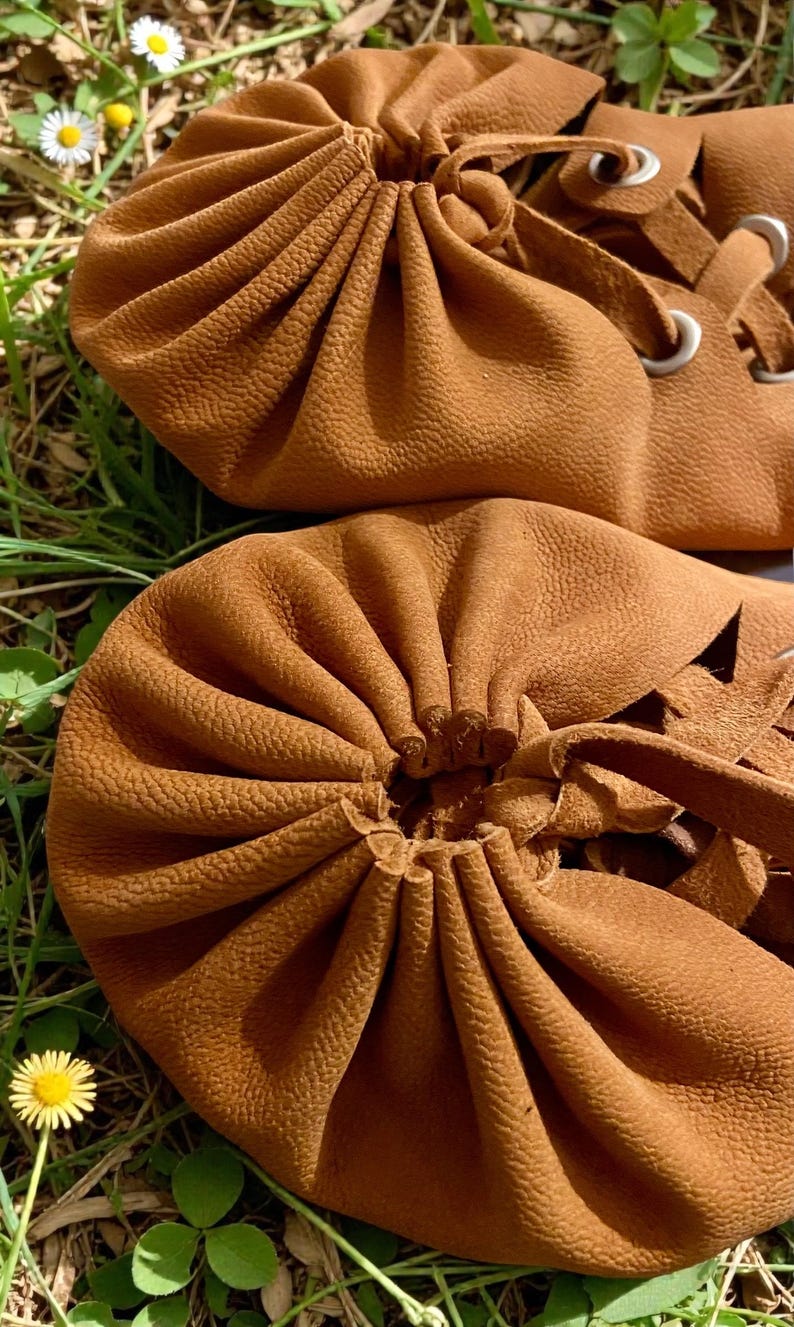 May include: Close-up of two brown leather drawstring pouches. The pouches have a textured surface and are gathered at the top with a leather tie. The background is green grass with small white and yellow flowers.