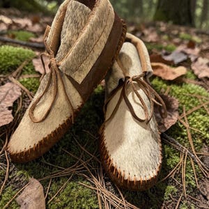 May include: A pair of handmade moccasins with a natural fur upper and brown leather accents. The shoes feature a soft, light-colored fur, dark brown leather soles, and leather laces. The moccasins are designed with a classic, traditional style.
