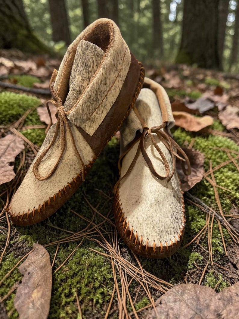 May include: A pair of handmade moccasins with a natural fur upper and brown leather accents. The shoes feature a soft, light-colored fur, dark brown leather soles, and leather laces. The moccasins are designed with a classic, traditional style.