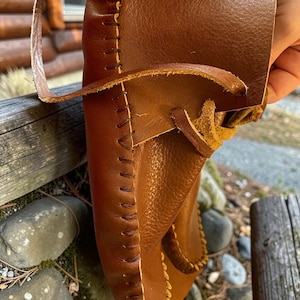 May include: A brown leather moccasin featuring a visible whipstitch seam along the edge and a leather lace threaded through the upper section. The shoe is constructed from a smooth, flexible material with a soft, unlined interior texture.