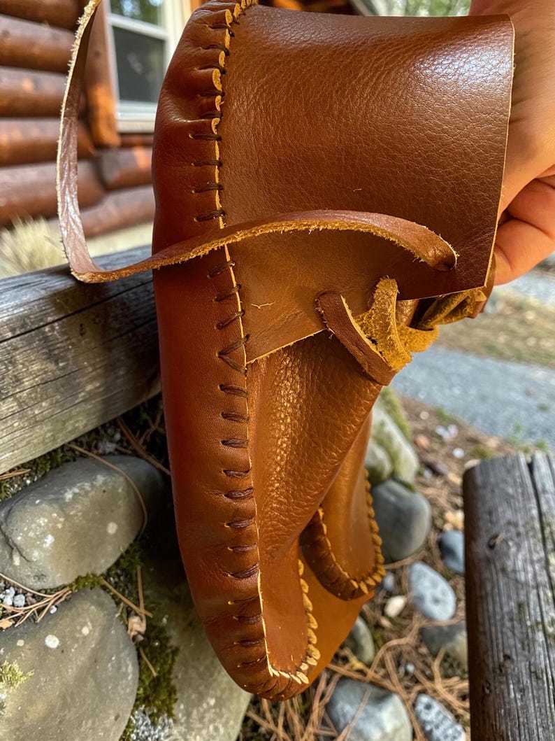 May include: A brown leather moccasin featuring a visible whipstitch seam along the edge and a leather lace threaded through the upper section. The shoe is constructed from a smooth, flexible material with a soft, unlined interior texture.