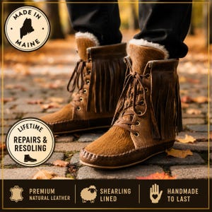 May include: Brown leather ankle boots with a textured hair-on-hide finish, featuring long fringe detailing, lace-up closures with metal eyelets, and a white shearling lining. The boots include text labels stating Made in Maine and Lifetime Repairs & Resoling.