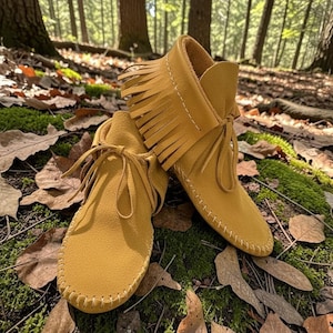 May include: Mustard yellow suede moccasin boots with fringe detailing and tie closures. The boots are shown outdoors on a bed of leaves and moss, with a forest background. The boots have a soft, flexible sole and are stitched with white thread.