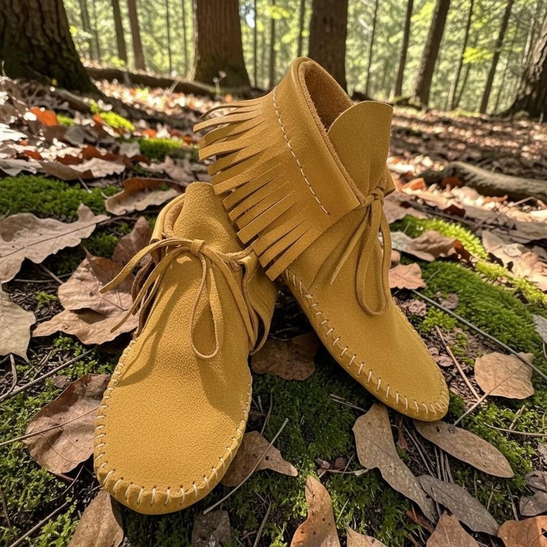 May include: Mustard yellow suede moccasin boots with fringe detailing and tie closures. The boots are shown outdoors on a bed of leaves and moss, with a forest background. The boots have a soft, flexible sole and are stitched with white thread.