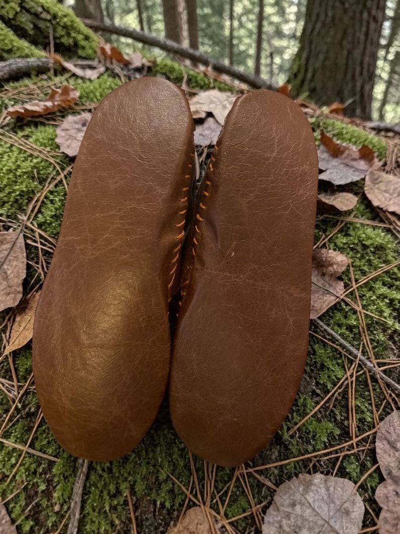 May include: A pair of brown leather moccasins, soles facing up, with visible stitching. The shoes are set against a backdrop of moss, leaves, and forest floor, suggesting an outdoor or natural theme.