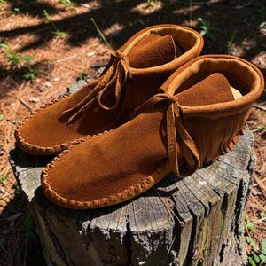Puede incluir: Un par de mocasines de ante color marr&oacute;n tabaco con una construcci&oacute;n suave y flexible, costuras visibles a lo largo de las punteras redondeadas, un cuello alto y detalles de flecos decorativos en los laterales con cordones finos de ante.