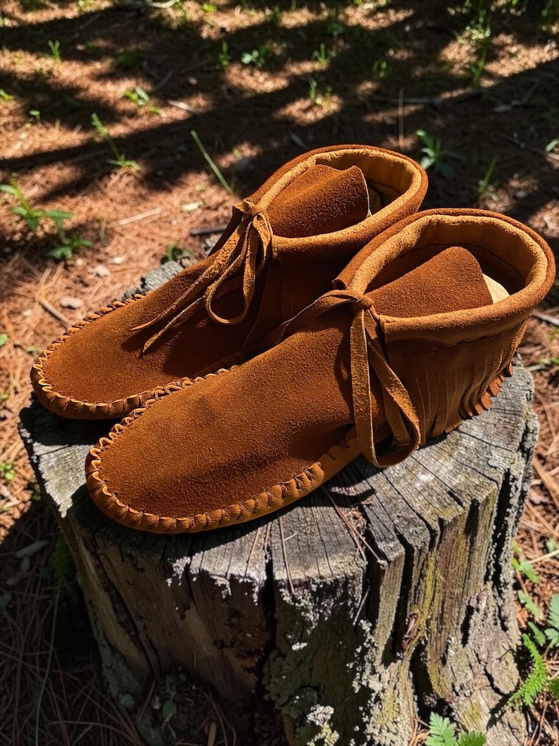 Puede incluir: Un par de mocasines de ante color marr&oacute;n tabaco con una construcci&oacute;n suave y flexible, costuras visibles a lo largo de las punteras redondeadas, un cuello alto y detalles de flecos decorativos en los laterales con cordones finos de ante.