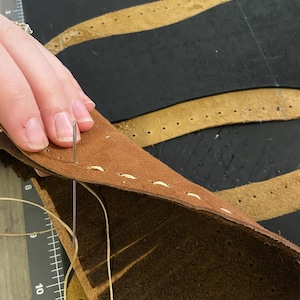 May include: Close-up of leather craft in progress. A needle and thread are used to stitch together two pieces of brown leather. The leather has pre-punched holes for stitching. The background shows additional leather pieces and a ruler.