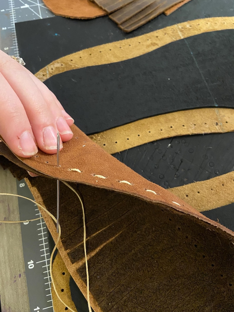 May include: Close-up of leather craft in progress. A needle and thread are used to stitch together two pieces of brown leather. The leather has pre-punched holes for stitching. The background shows additional leather pieces and a ruler.