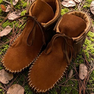 Puede incluir: Un par de mocasines de ante marr&oacute;n hasta el tobillo con un forro interior suave y difuso, detalles de flecos decorativos en la parte posterior y una construcci&oacute;n de costura visible alrededor de la puntera con cordones de cuero.
