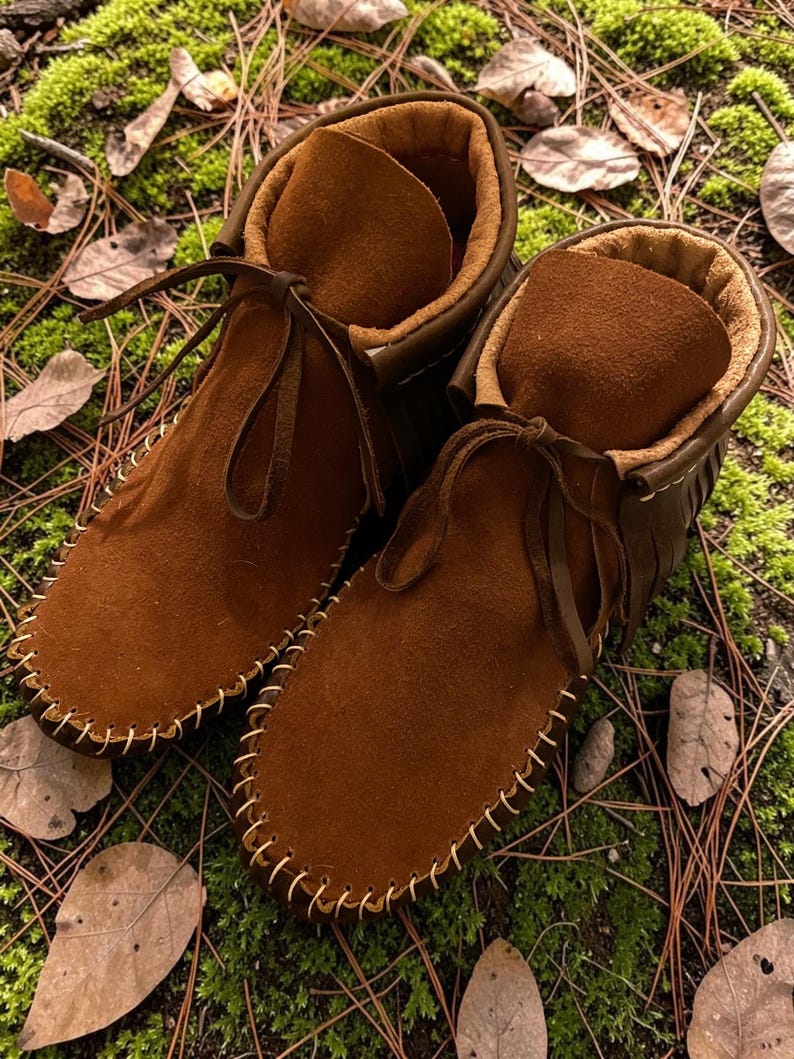 Puede incluir: Un par de mocasines de ante marr&oacute;n hasta el tobillo con un forro interior suave y difuso, detalles de flecos decorativos en la parte posterior y una construcci&oacute;n de costura visible alrededor de la puntera con cordones de cuero.