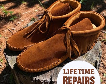 Puede incluir: Botas mocasín de ante marrón con cordones de cuero y flecos. Se muestran sobre un tocón de árbol. La imagen incluye un círculo blanco con el texto "LIFETIME REPAIRS & RESOLING!"