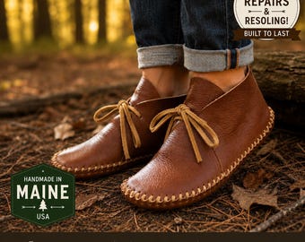Può includere: Stivaletti mocassino in pelle marrone con cuciture a vista lungo la suola e lacci in pelle color cuoio chiaro. Gli stivali sono indossati con jeans blu scuro con risvolto. Il testo sull'immagine include "LIFETIME REPAIRS & RESOLING!" e "HANDMADE IN MAINE USA".